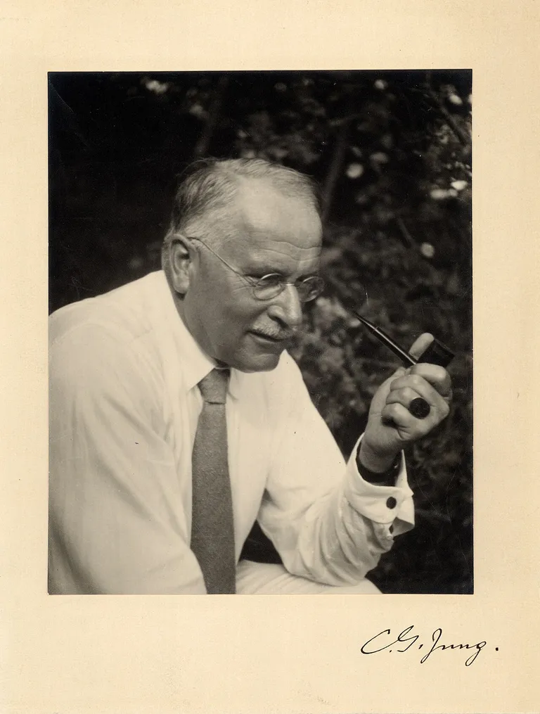 Jung in his sixties, in shirtsleeves and a tie, seated outdoors and turned toward the camera with a pipe raised in one hand. The mount is signed C. G. Jung.